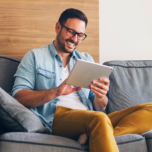 Man with glasses on couch using tablet and smiling