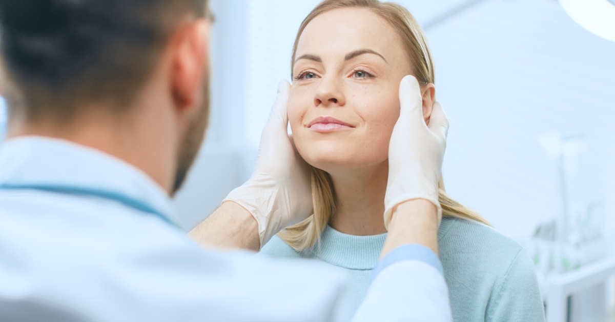 A cosmetic surgeon examines a beautiful woman's face with gloved hands while sitting in a clinic room.